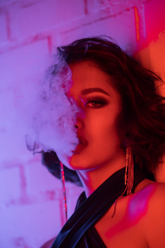 Retrato de mujer asiática joven de moda con maquillaje exhalando humo en luz de neón en club nocturno - Foto, imagen