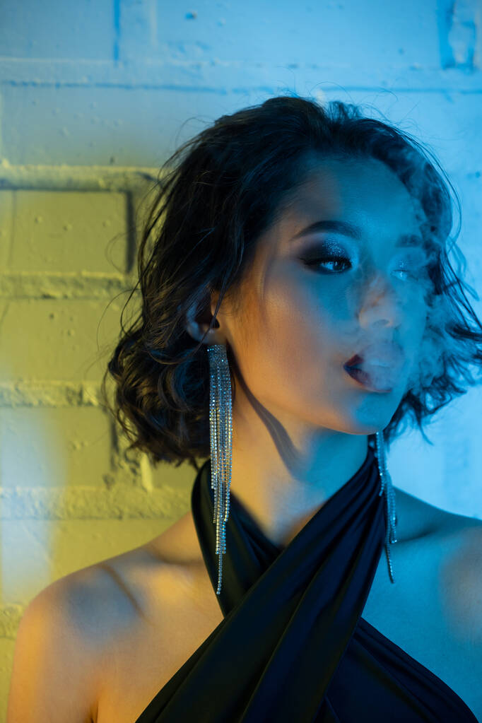 Portrét módní mladé asijské ženy s make-upem vydechující kouř v neonovém světle v nočním klubu - Fotografie, Obrázek