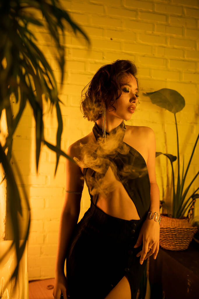 Gece kulübünde sarı ışıkta bitkilerin yanında duran seksi Asyalı kadın. - Fotoğraf, Görsel
