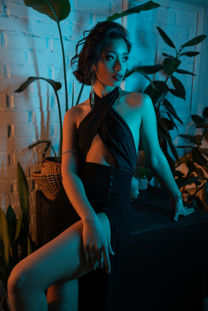 Elegante joven asiática mujer en vestido mirando lejos cerca de plantas en luz de neón en club nocturno - Foto, Imagen
