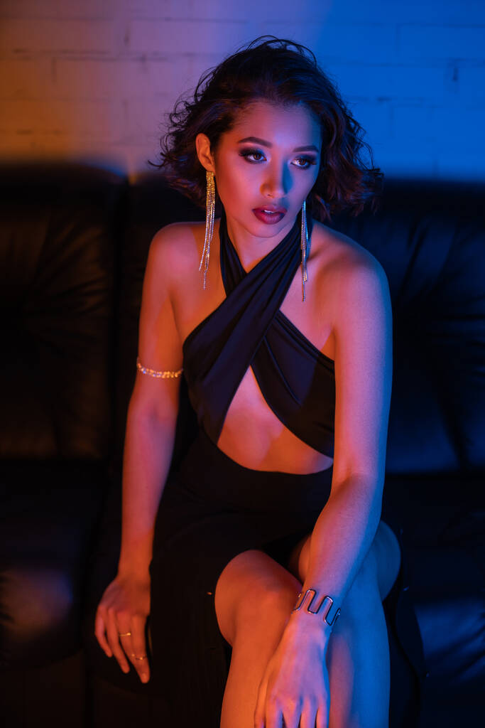 Модна азіатка в сережках, браслетах і сукні сидить на дивані з неоновим світлом в нічному клубі - Фото, зображення