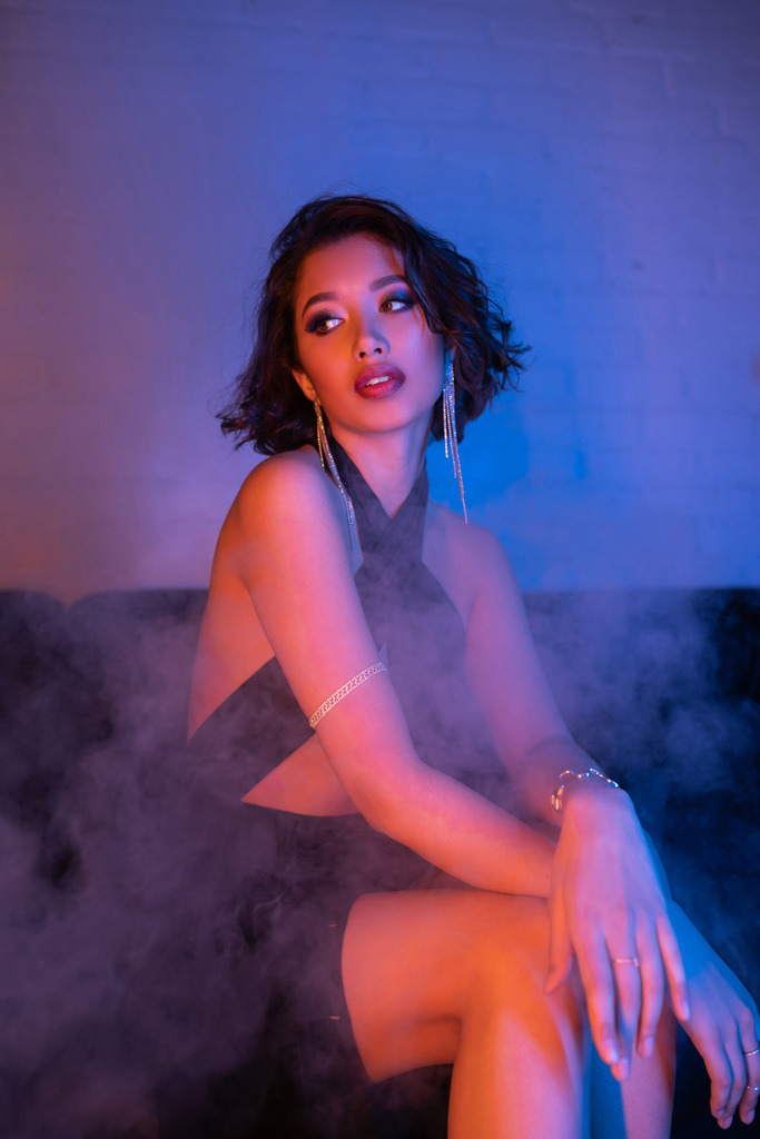 modne młody Azji kobieta w sukienka siedzi w dym i neon światło na kanapie w nocny klub - Zdjęcie, obraz