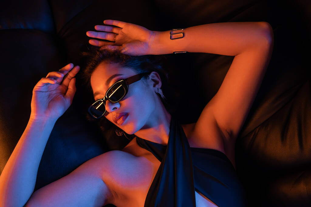 Widok z góry młodej Azjatki w okularach przeciwsłonecznych i sukience leżącej na kanapie w neonowym świetle w klubie nocnym - Zdjęcie, obraz