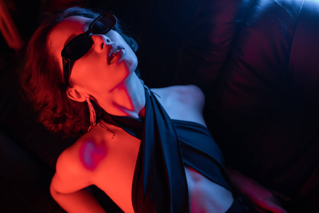 High angle view of sexy asian woman in sunglasses posing in neon light on couch in night club - Photo, Image