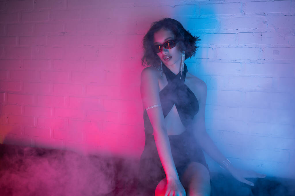 Młoda Azjatka w okularach przeciwsłonecznych siedzi na kanapie podczas imprezy z dymem i neonem w klubie nocnym - Zdjęcie, obraz