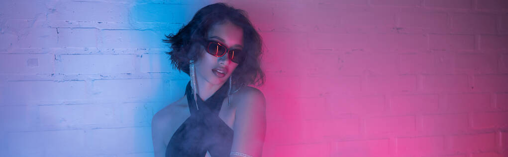 Młoda Azjatka w okularach przeciwsłonecznych pozująca w dymie i neonowym oświetleniu w klubie nocnym, sztandar - Zdjęcie, obraz
