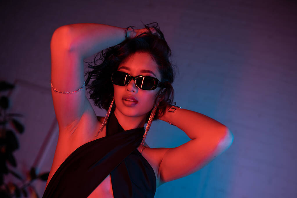 Модная азиатка в солнечных очках и платье трогательная голова в красочном неоновом свете в ночном клубе - Фото, изображение