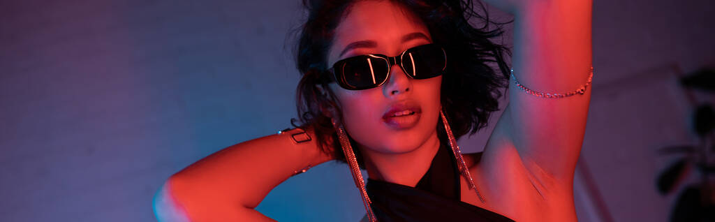 Güneş gözlüklü ve bilezikli modaya uyan Asyalı kadın gece kulübünde renkli neon ışıkta poz veriyor. - Fotoğraf, Görsel