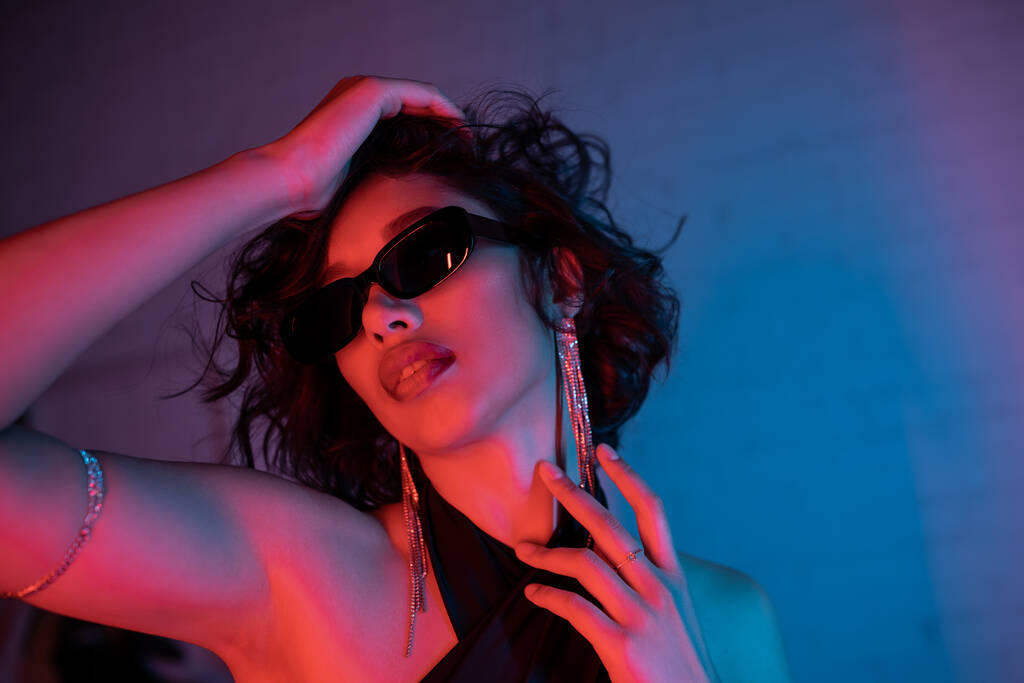 Joven mujer asiática en gafas de sol tocando la cabeza mientras está de pie en la luz de neón colorido en el club nocturno - Foto, imagen