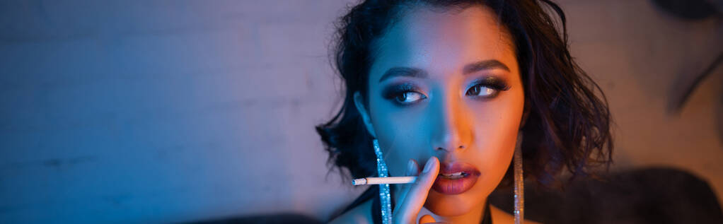 Fashionable asian woman with makeup and hairstyle smoking cigarette in night club, banner - Photo, Image