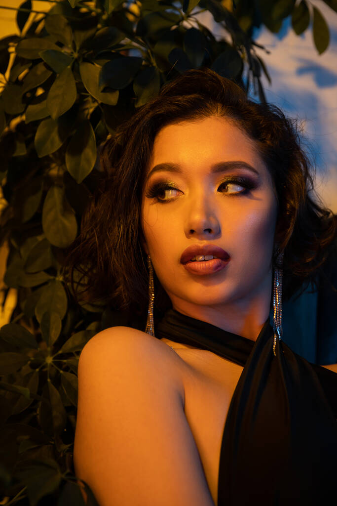 Confiado joven mujer asiática con maquillaje mirando lejos cerca de la planta en el club nocturno con iluminación - Foto, imagen