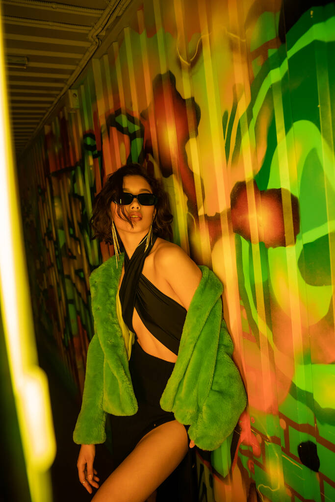 sexy asiatico donna in occhiali da sole e falso pelliccia giacca in piedi vicino graffiti in notte club - Foto, immagini
