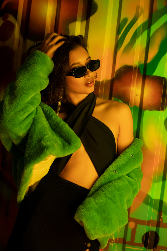 Модна молода азіатка в сонцезахисних окулярах торкається голови біля графіті на стіні в нічному клубі - Фото, зображення