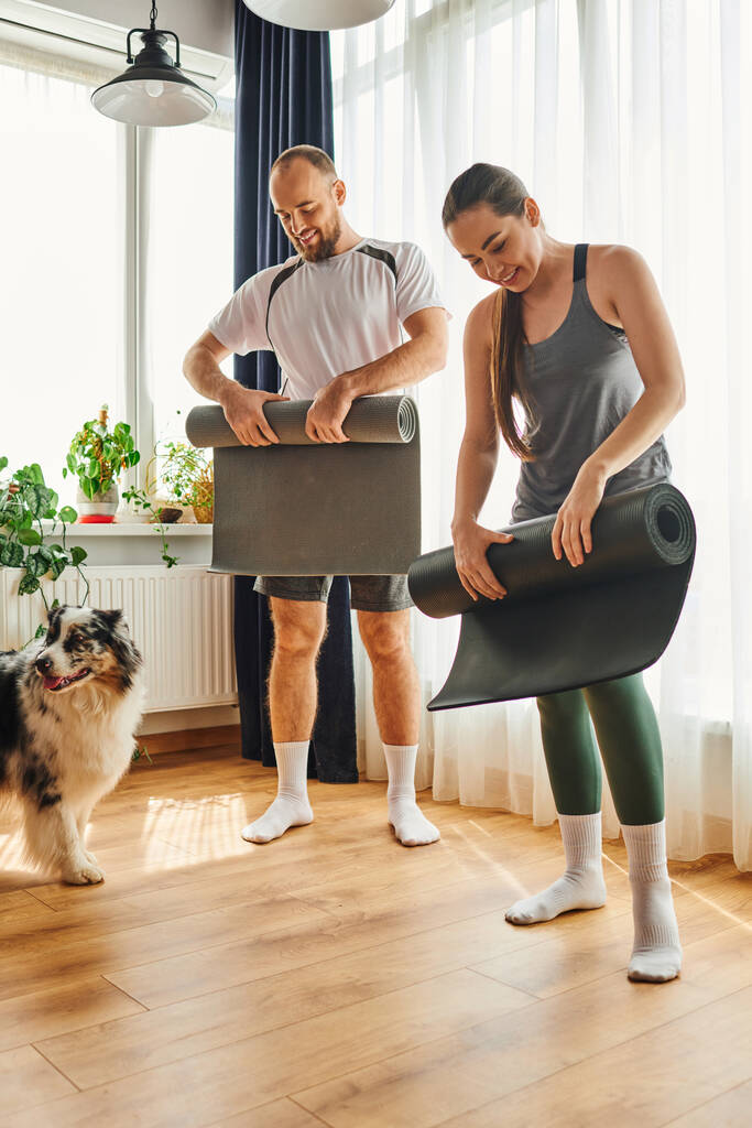 Cheerful couple in sportswear holding fitness mats near border collie in living room at home - Photo, Image