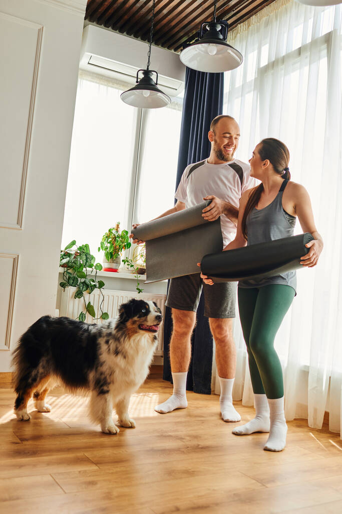 Positive couple in sportswear with fitness mats looking at each other near border collie at home - Photo, Image