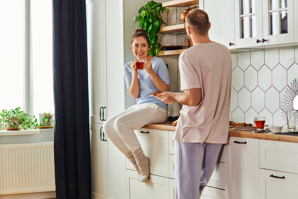 Uśmiechnięta kobieta w homewear trzymając herbatę w pobliżu chłopaka ze śniadaniem na talerzu w kuchni rano - Zdjęcie, obraz