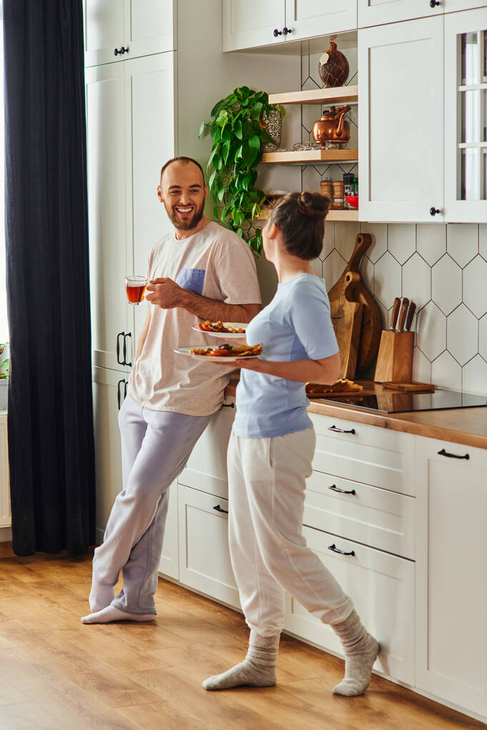 Veselý vousatý muž v domácím oblečení drží čaj a dívá se na přítelkyni s preakfast v kuchyni - Fotografie, Obrázek