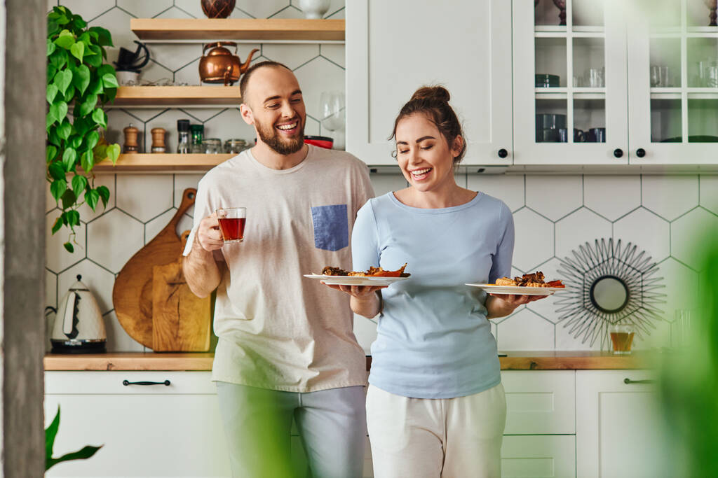 Hymyilevä nainen homewear tilalla levyt aamiaisella lähellä poikaystävä teetä keittiössä - Valokuva, kuva