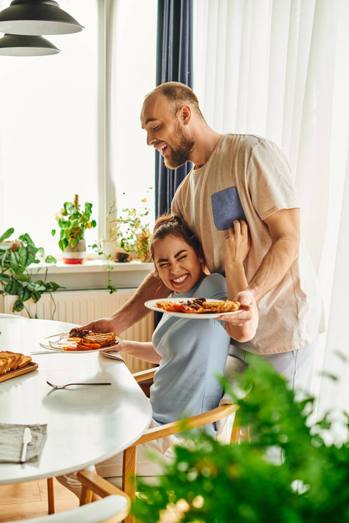 Femme brune positive touchant petit ami mettant savoureux petit déjeuner sur la table à la maison le matin - Photo, image