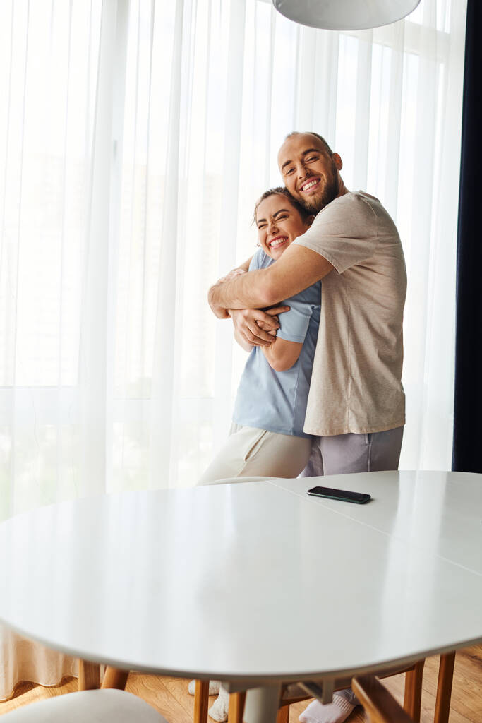 Uśmiechnięty brodaty mężczyzna w homewear przytulając dziewczynę w pobliżu smartfona na stole w domu - Zdjęcie, obraz