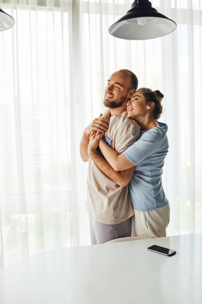 Hymyilevä nainen homewear halaus poikaystävä ja seisoo lähellä älypuhelin tyhjä näyttö kotona - Valokuva, kuva