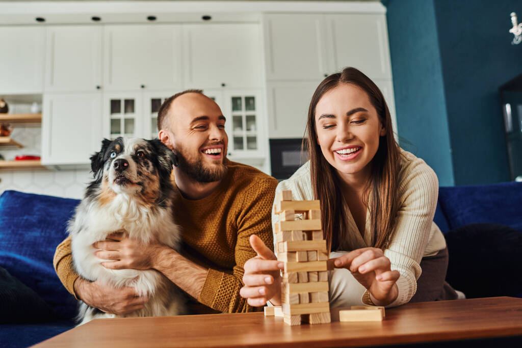 Femme souriante jouant jeu de blocs de bois près du petit ami et collie frontière dans le salon à la maison - Photo, image