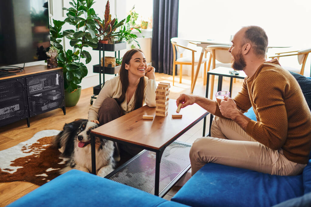 Uomo sorridente in possesso di caffè e giocare blocchi di legno gioco con la ragazza vicino al confine collie a casa - Foto, immagini