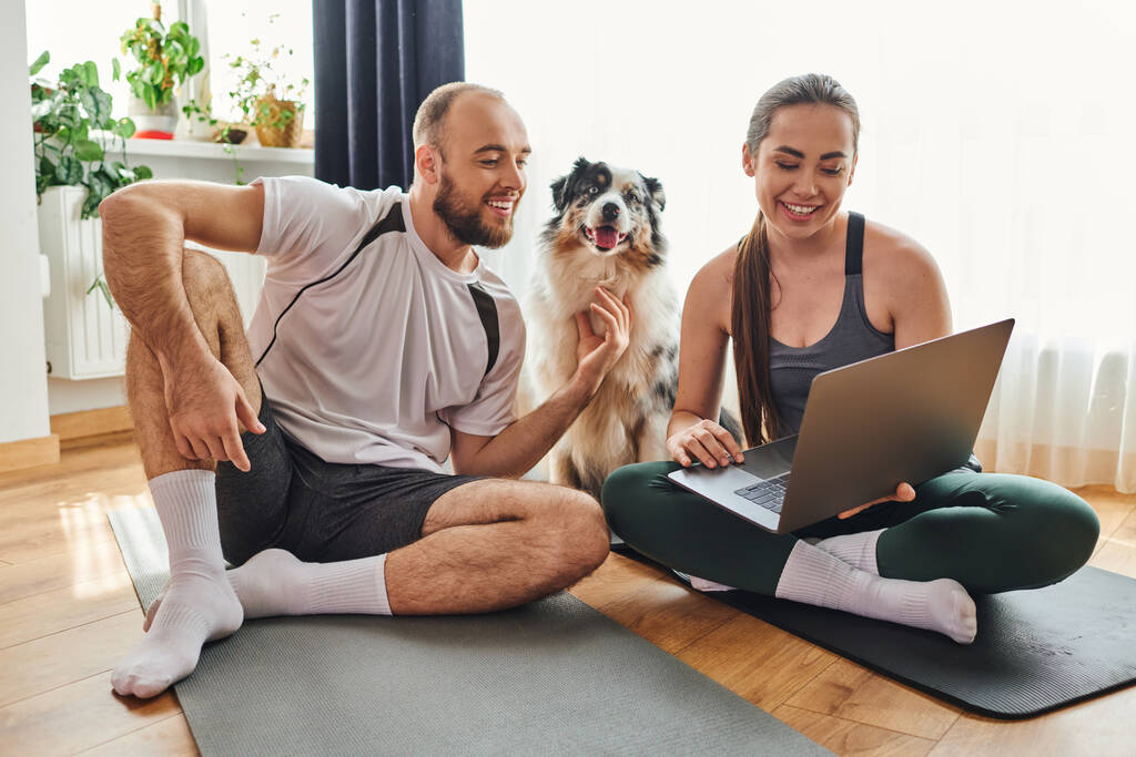 Smiling woman using laptop and sitting on fitness mat near boyfriend and border collie at home - Photo, Image