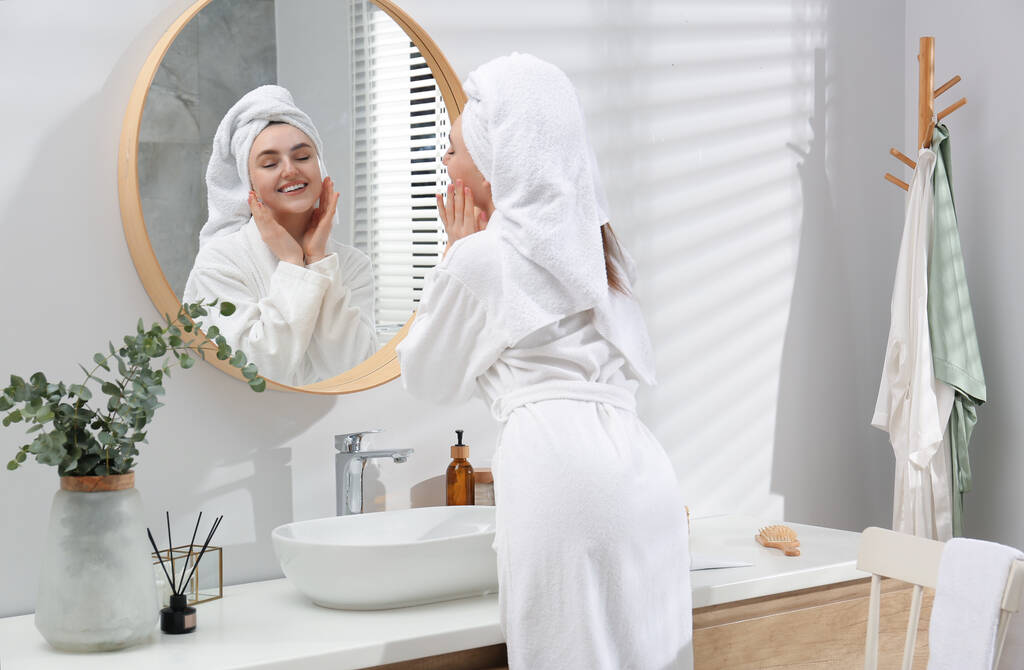 Kaunis nainen valkoisessa kaapussa soveltamalla kerma lähellä peiliä kylpyhuoneessa - Valokuva, kuva