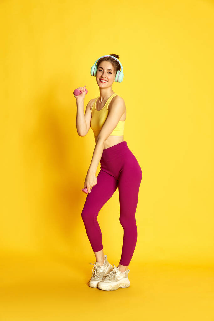 Full-length image of young slim woman in sportswear and hedges, training with dumbbells against yellow studio background. Concept of sport, fitness, body care, fashion, youth, lifestyle, ad - Photo, Image