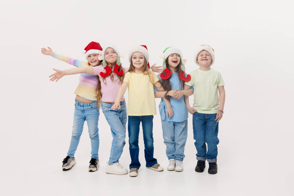 С Новым годом и Рождеством! Группа веселых счастливых детей в праздничном Санта-Клаусе и эльфийских шляпах на белом фоне студии. Дети обнимаются и веселятся - Фото, изображение