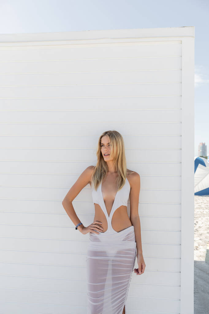 Een jonge blonde vrouw in een vloeiende witte jurk poseert sierlijk naast een grimmige witte muur in een Miami strand setting. - Foto, afbeelding