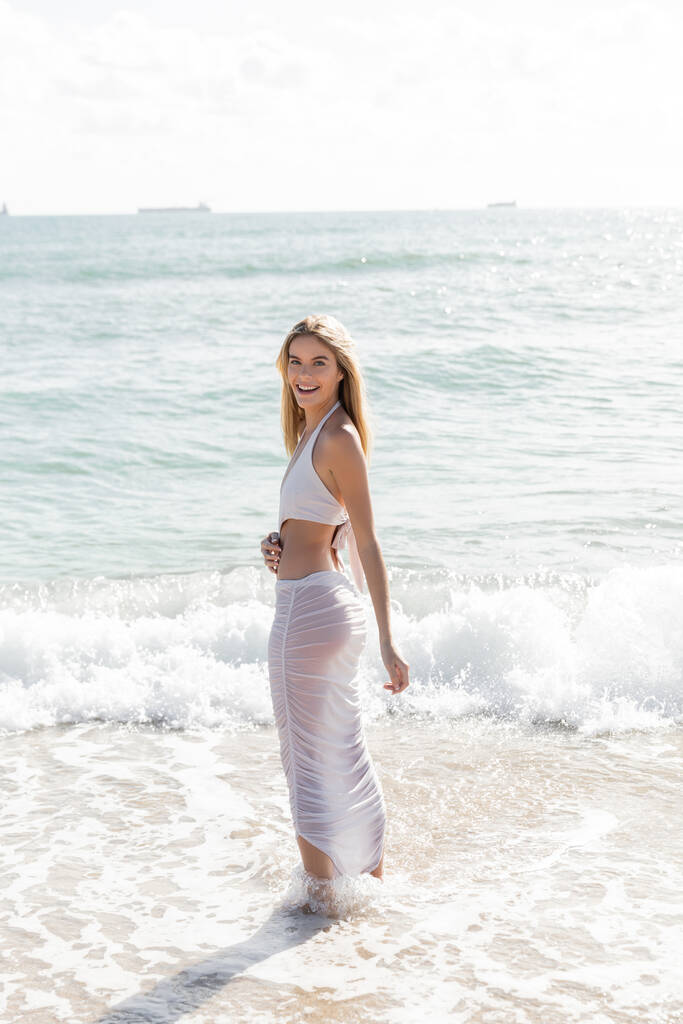 Een jonge, mooie blonde vrouw staat gracieus in het water op Miami Beach op een zonnige dag. - Foto, afbeelding