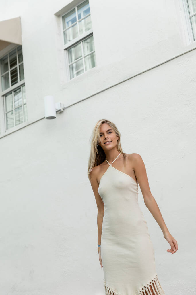 Uma jovem mulher loira em um vestido branco fluindo posa elegantemente na frente de um magnífico edifício em Miami. - Foto, Imagem