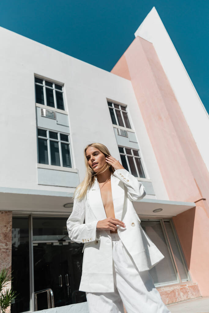 Upea blondi nainen valkoisessa puvussa poseeraa suuren rakennuksen edessä Miamissa.. - Valokuva, kuva