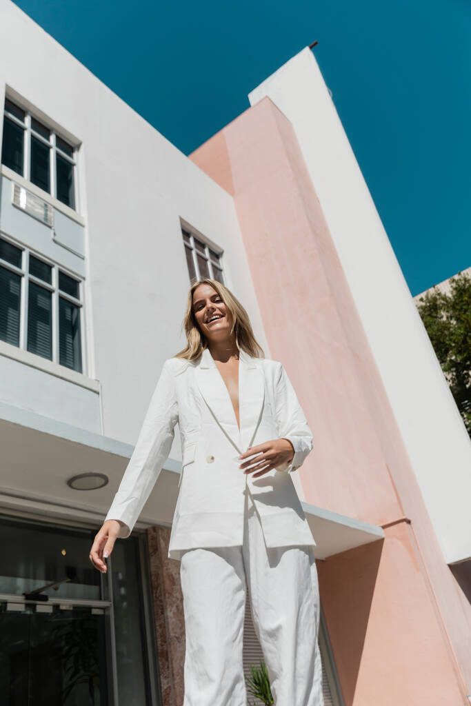 Una giovane, bella donna bionda si trova con fiducia in un abito bianco di fronte a uno splendido edificio di Miami. - Foto, immagini