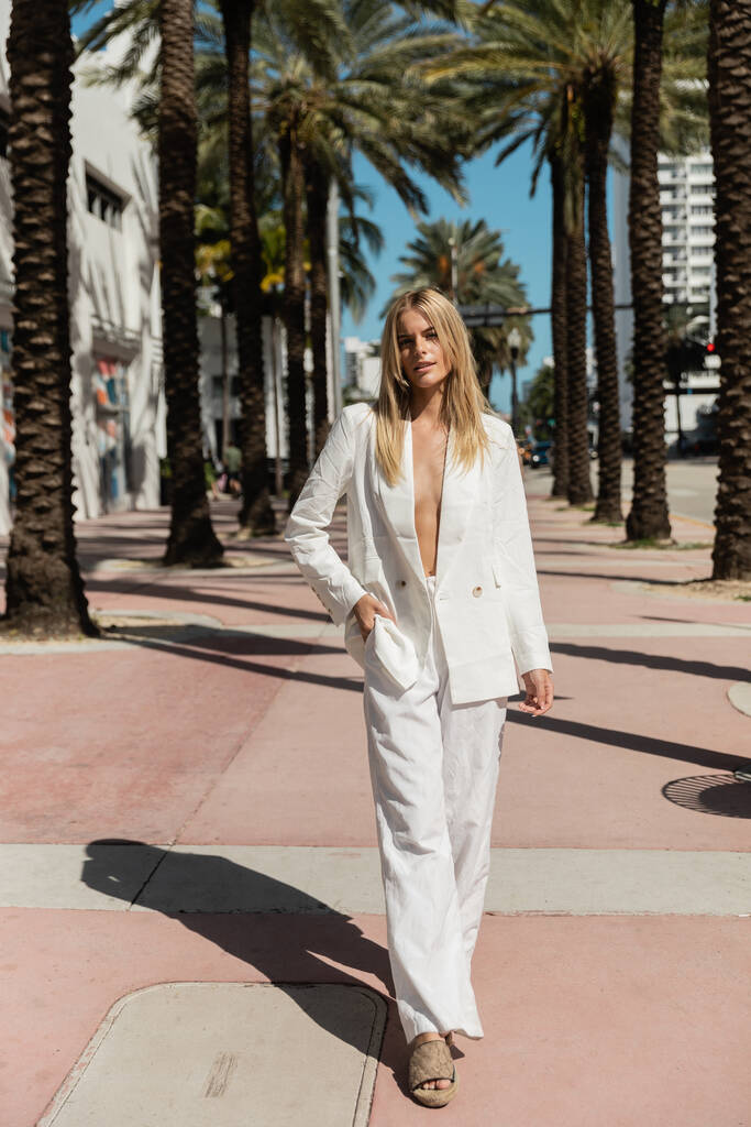 Une femme blonde respire la confiance alors qu'elle descend une rue de Miami en costume blanc époustouflant, une vision d'élégance. - Photo, image