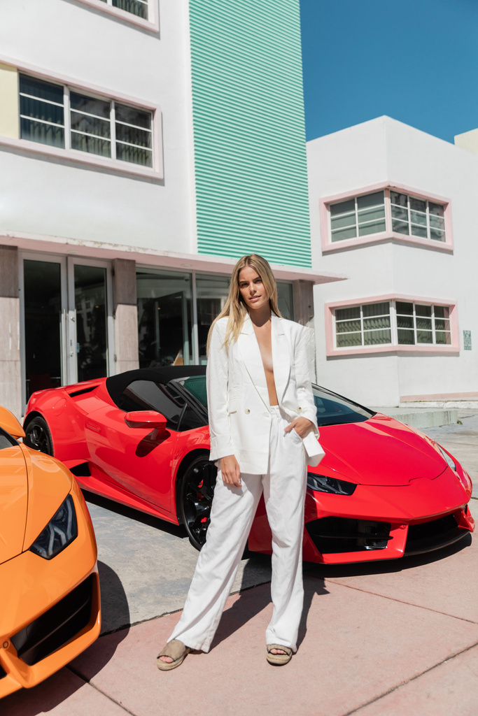 A young blonde woman standing confidently in front of two sleek sports cars. - Photo, Image
