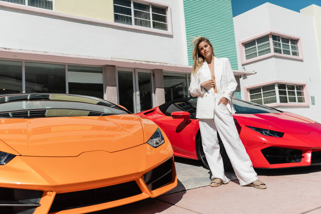 Молода блондинка впевнено стоїть поруч з двома гладкими спортивними автомобілями в Маямі. - Фото, зображення