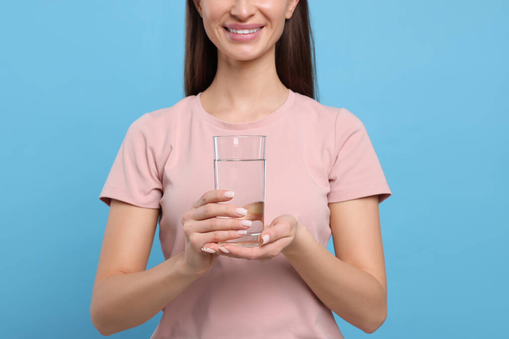 Un hábito saludable. Mujer feliz sosteniendo vidrio con agua dulce sobre fondo azul claro, primer plano - Foto, Imagen