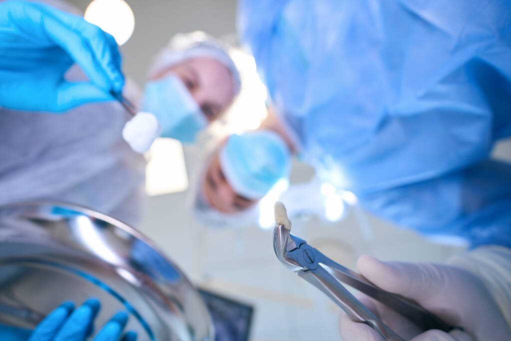 Чоловічий хірург тримає щипці з видаленим зубом в руках, молодий асистент має пінцет з тампоном в руках - Фото, зображення