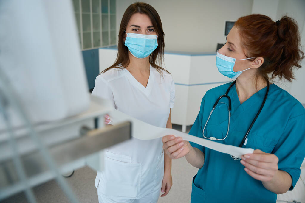 Медсестра и врач общей практики в медицинских масках обсуждают анализ пациента после предыдущего обследования в стационаре - Фото, изображение
