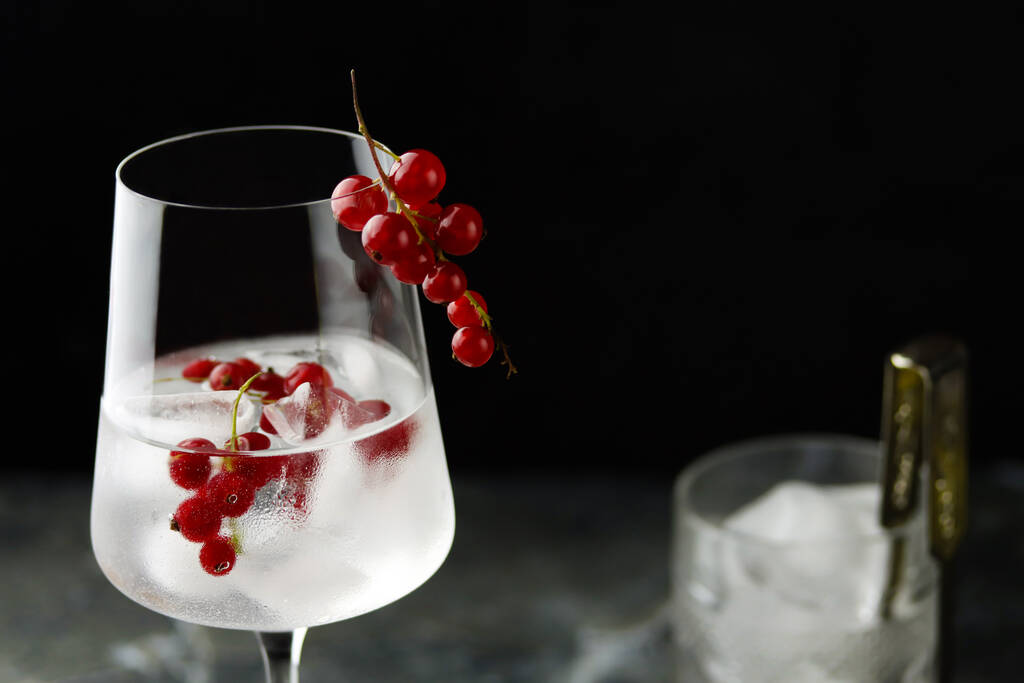 Ekstremalne zbliżenie kieliszka do wina z mrożoną wodą z czerwonej porzeczki. Świeży koktajl owocowy w szkle, orzeźwiający letni napój jagodowy z porzeczki na ciemnym tle, widok z przodu. Picie z czerwonej porzeczki - Zdjęcie, obraz