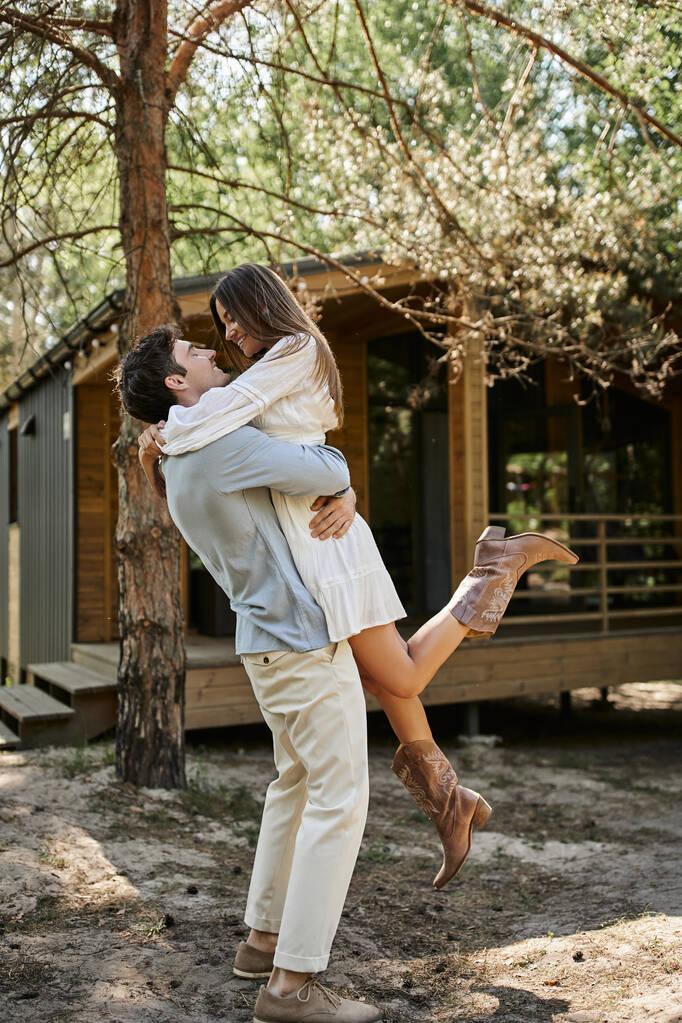 счастливый мужчина поднимает и обнимает женщину в доме отдыха, лесу, смотрит друг на друга, романтика и любовь - Фото, изображение