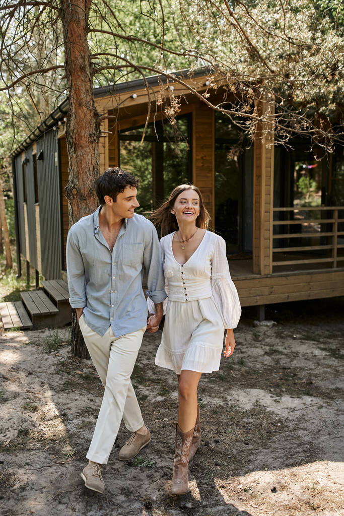 ευτυχισμένη γυναίκα κρατώντας τα χέρια και περπατώντας με χαμογελαστό φίλο, ζευγάρι κοντά στο σπίτι του καλοκαιριού, ρομαντισμός - Φωτογραφία, εικόνα