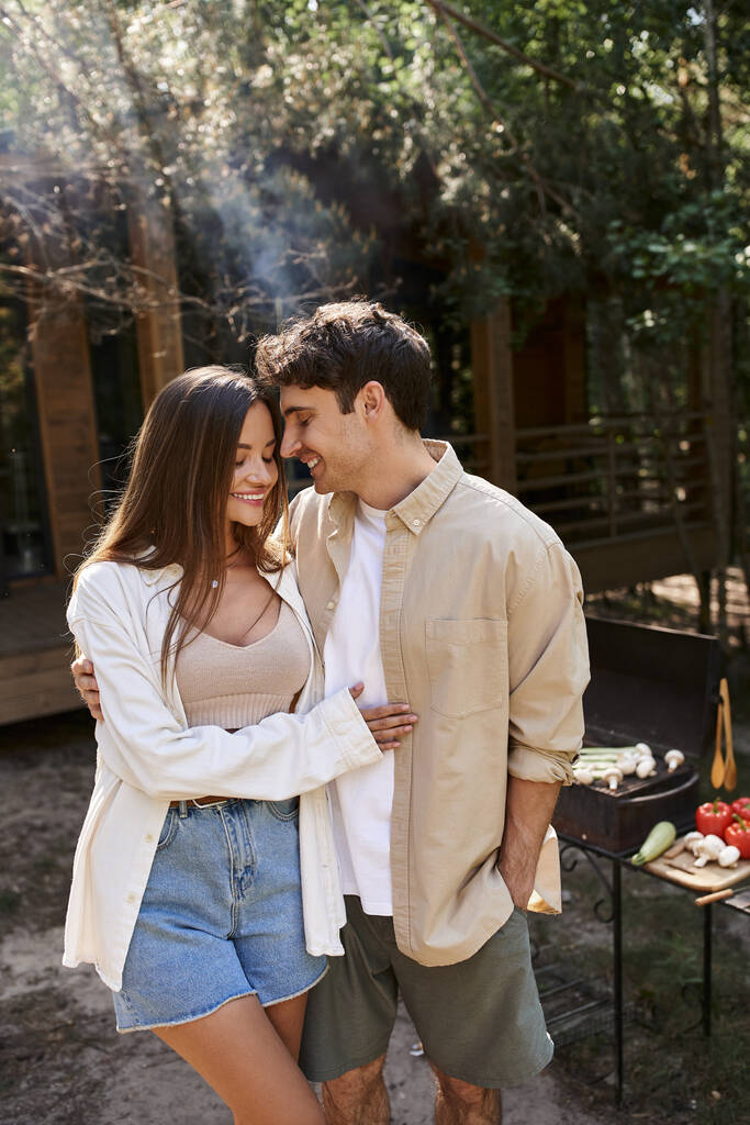 Hymyilevä romanttinen pari halaa lähellä hämärtynyt grilli ja loma talo taustalla ulkona - Valokuva, kuva