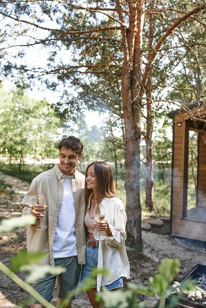 Χαρούμενο ζευγάρι με casual ρούχα που κρατάει κρασί κοντά στο μπάρμπεκιου και θολή εξοχική κατοικία στο παρασκήνιο - Φωτογραφία, εικόνα
