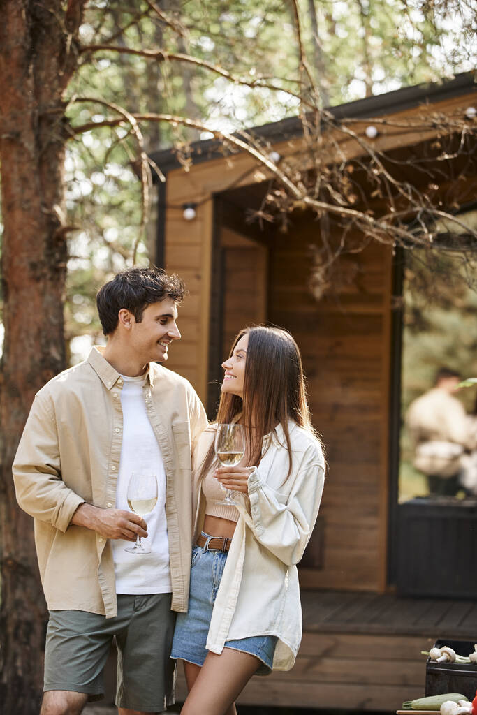 Positief brunette paar houden wijn tijdens de picknick in de buurt van barbecue en vakantiehuis op de achtergrond - Foto, afbeelding