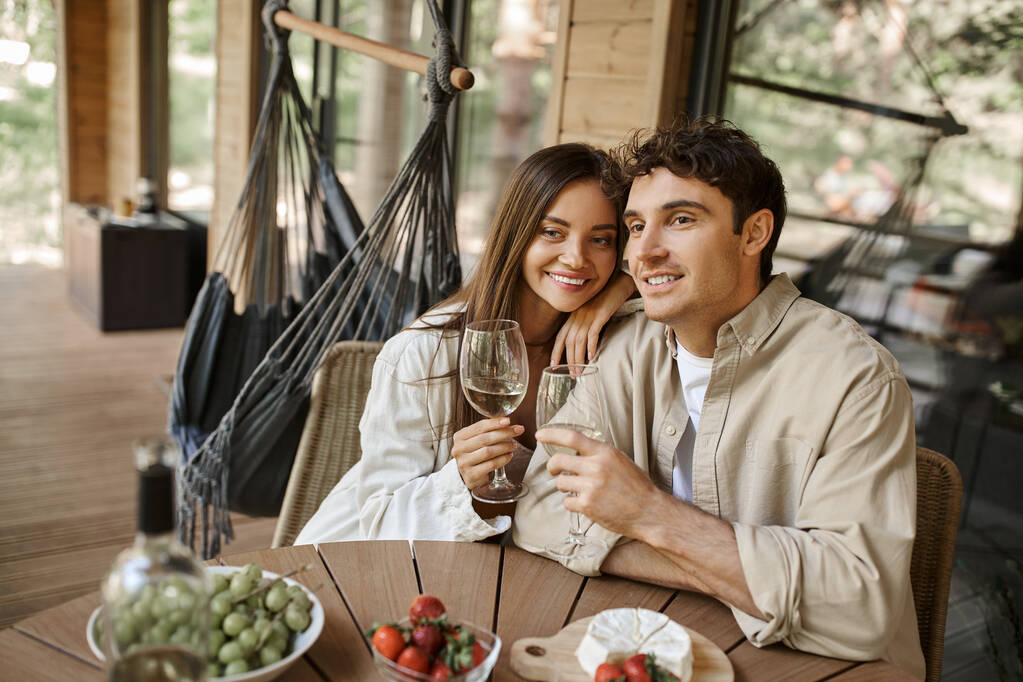 Счастливая романтическая пара, держащая вино рядом с фруктами и сыром на террасе дома отдыха летом - Фото, изображение
