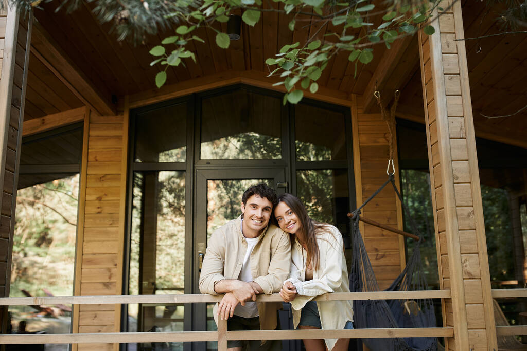 wesoła brunetka para patrząc w kamerę stojąc na tarasie drewnianego letniego domku na świeżym powietrzu - Zdjęcie, obraz